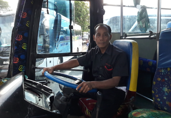 Kapudin dengan mengenakan seragam, tengah duduk di `kokpit` bus PO Dewi Sri yang dia kemudikan. 